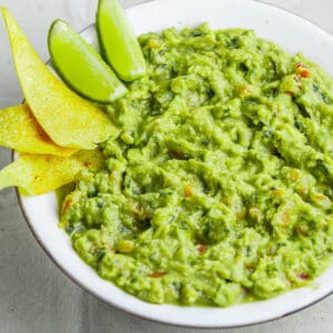 green guacamole in white bowl with lime and tortilla chips
