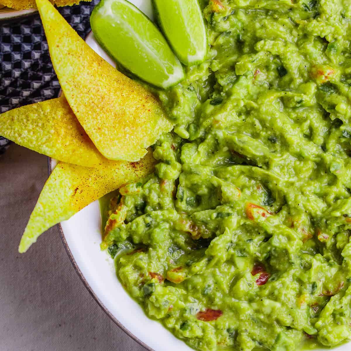 green guacamole in white bowl with lime and tortilla chips