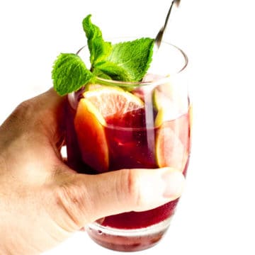 hand holding a glass of red sangria with mint, lemon, apples and ice