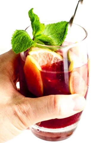 hand holding a glass of red sangria with mint, lemon, apples and ice