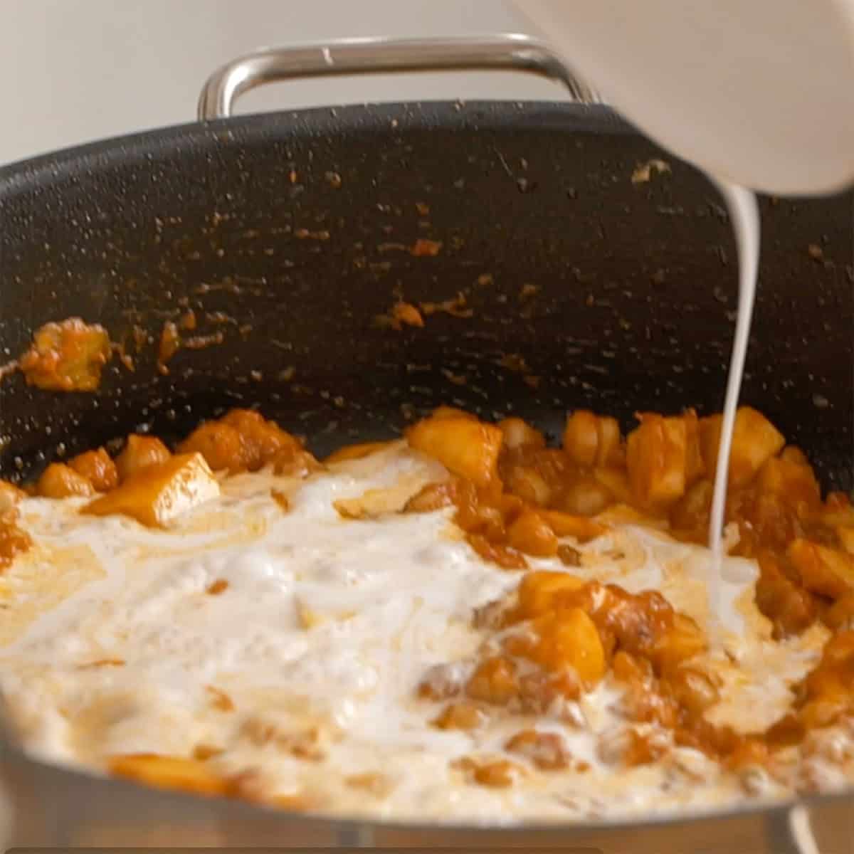 Adding coconut milk to curry in black pan.