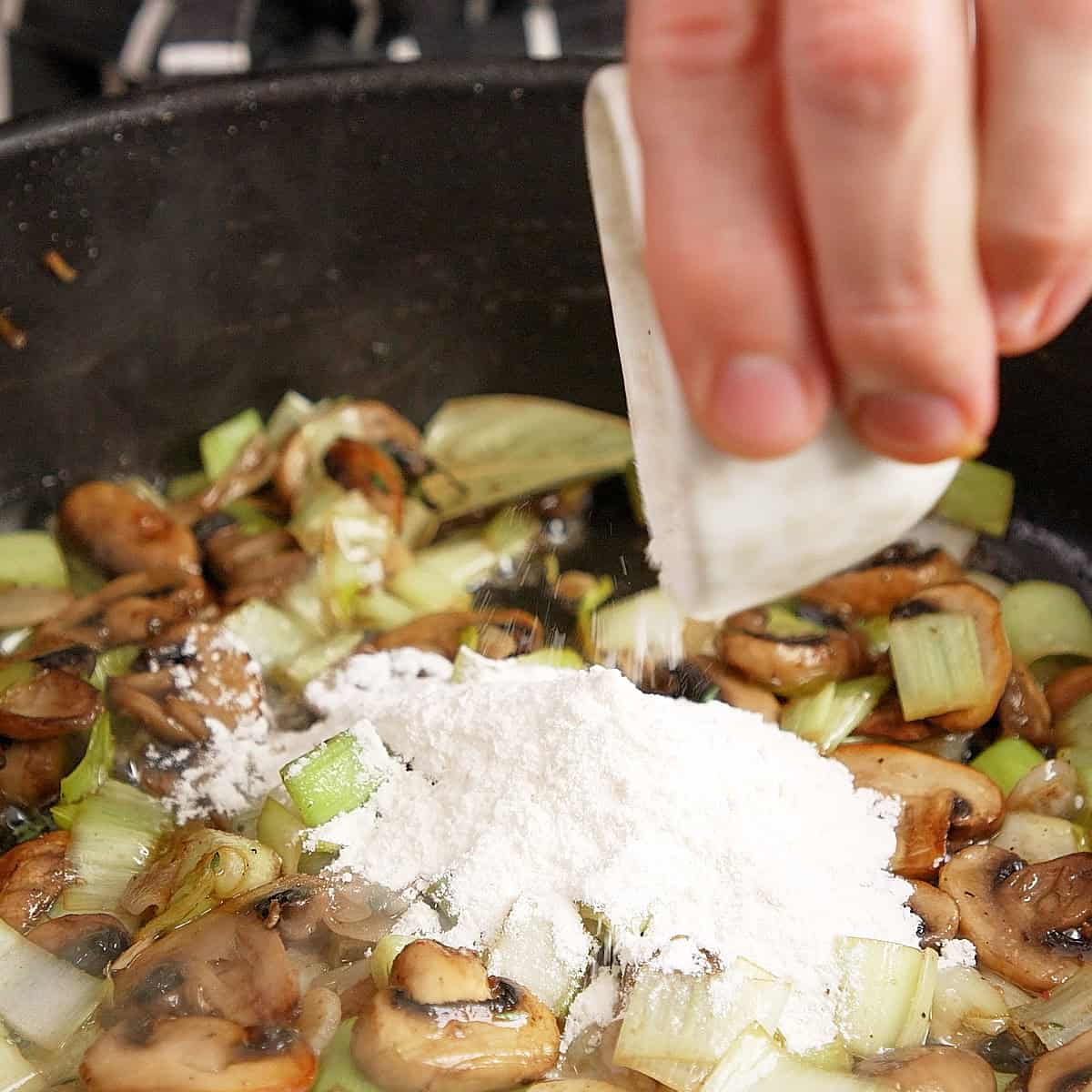 Adding flour to cooked out leeks and mushrooms in a black pan.