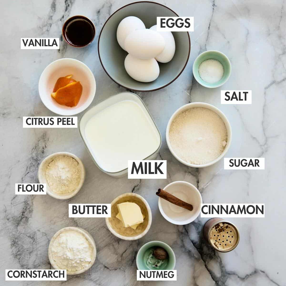 Three eggs, milk, sugar, flour, butter, cinnamon stick, and apricot jam arranged in small bowls on marble.