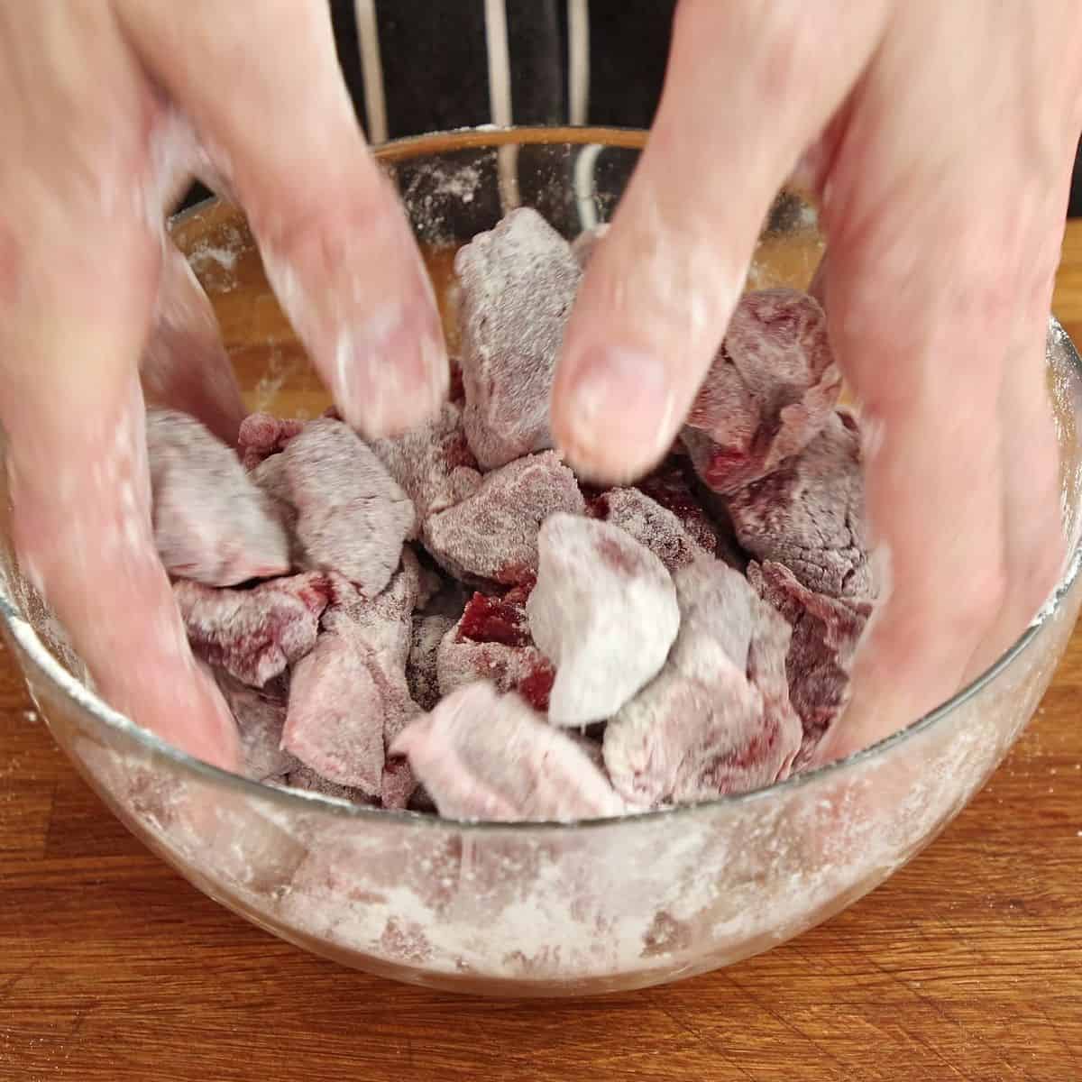 Beef chunks tossed with flour in metal bowl, flour coating visible on all pieces