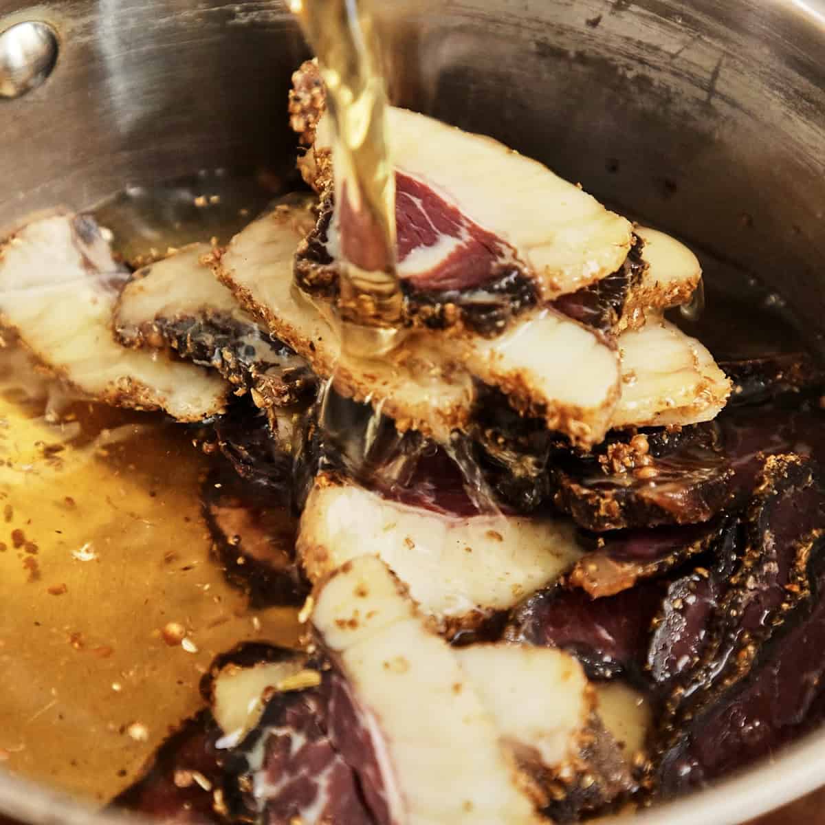 Sliced biltong steeping in warm beef stock in a stainless steel pot.