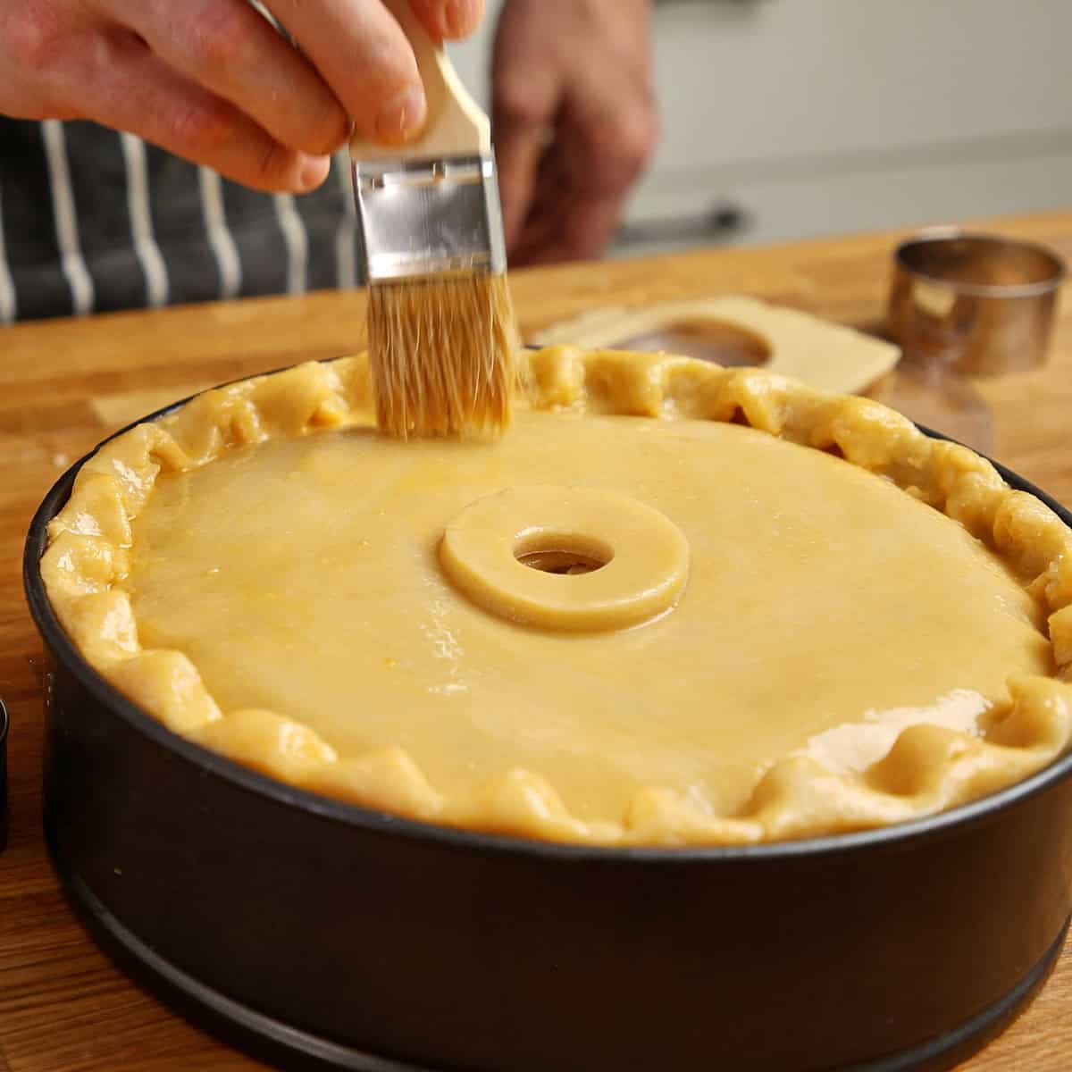 The pastry lid being brushed with egg wash, central steam hole visible.