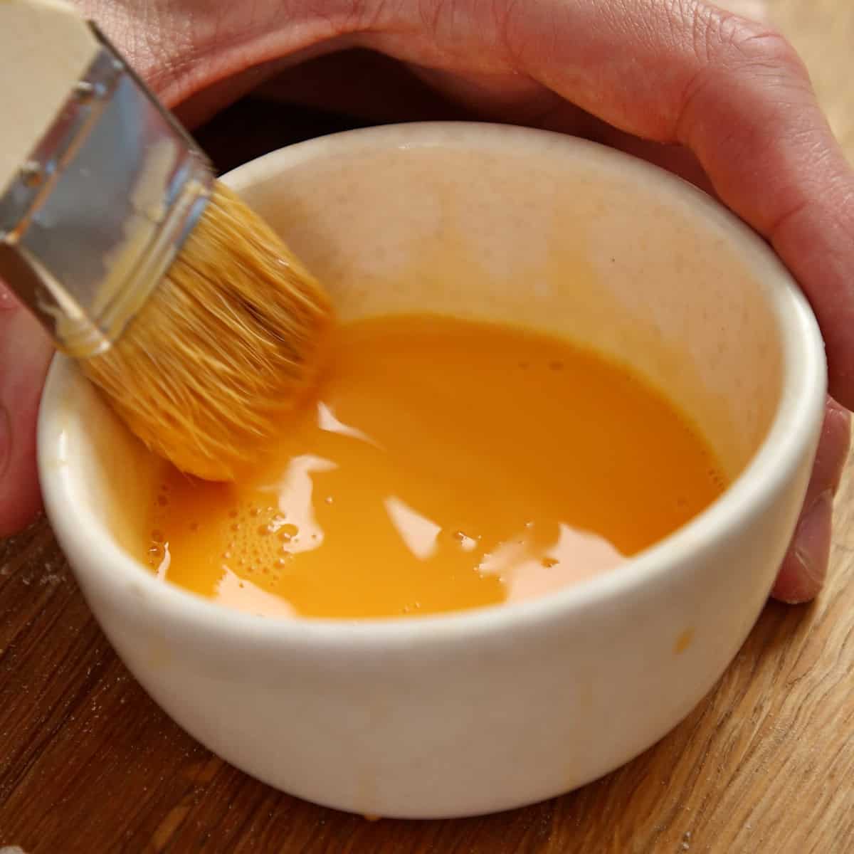 Egg wash being mixed in a white bowl with a pastry brush.