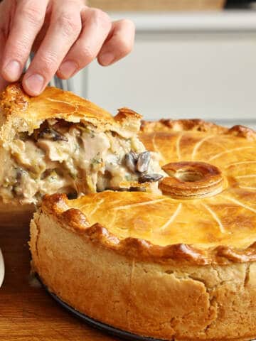Hand lifting a slice of baked chicken pie, revealing creamy filling with mushrooms and pastry crust.