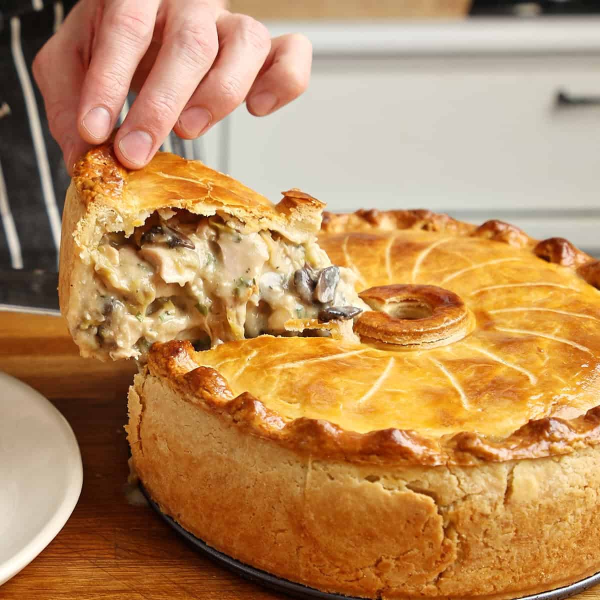 Hand lifting a slice of baked chicken pie, revealing creamy filling with mushrooms and pastry crust.