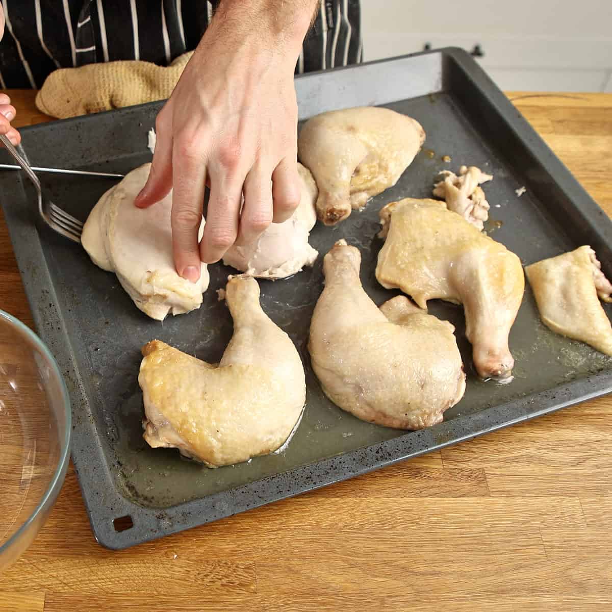 Roasted chicken being shredded with a fork on a dark baking tray.