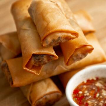 Stack of crispy spring rolls with dipping sauce on a wooden board.