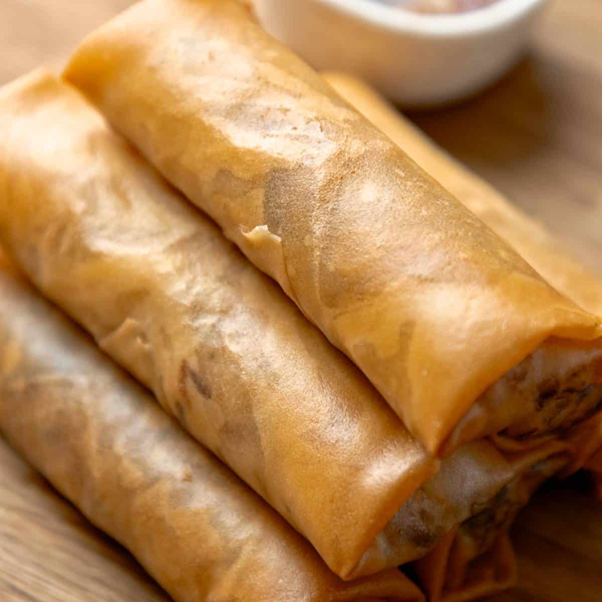 Stack of crispy spring rolls with dipping sauce on a wooden board.