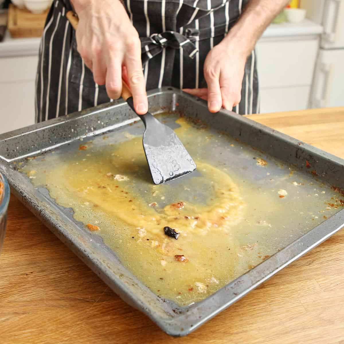 Hands scraping fond from a roasting tray with a metal spatula while deglazing.