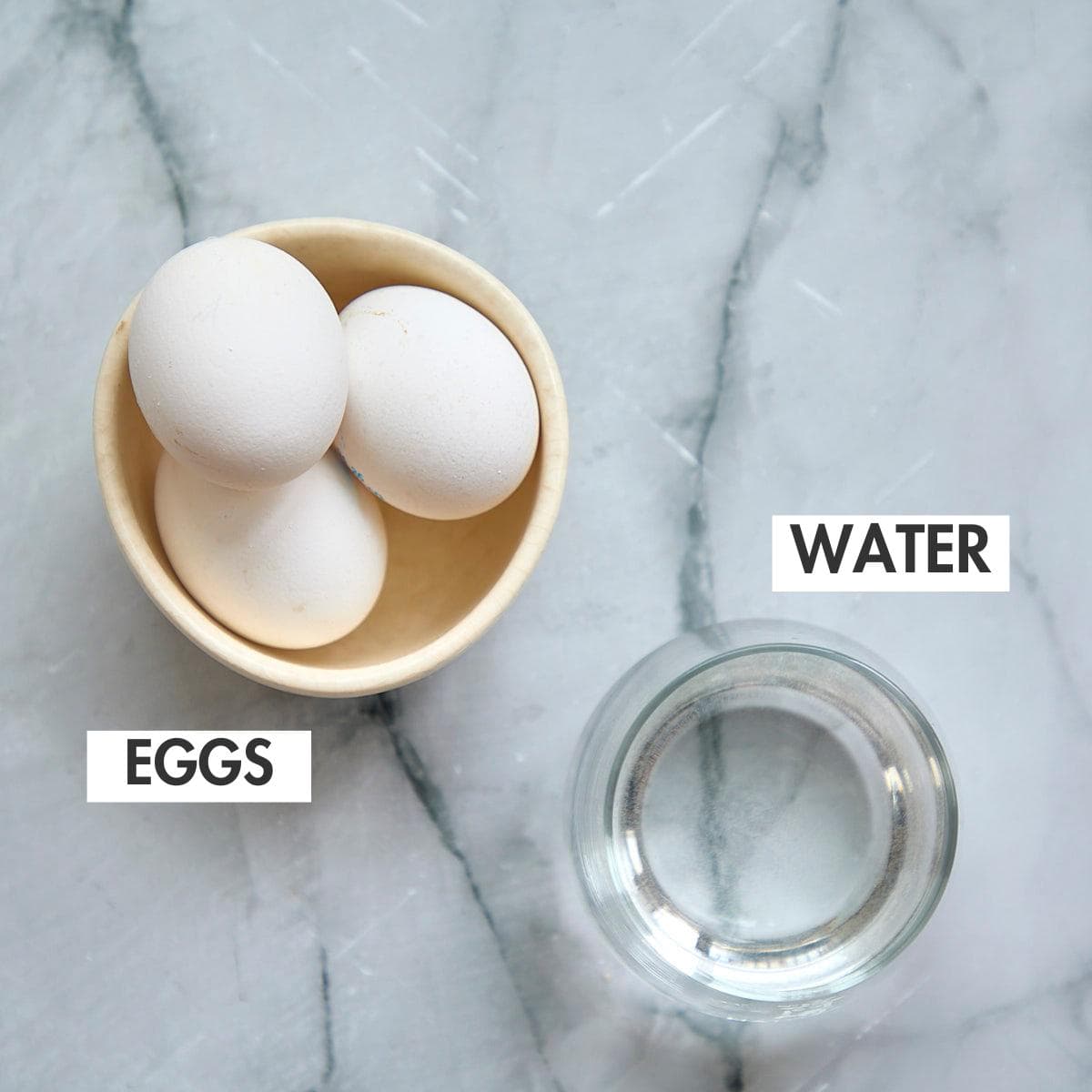 Three raw eggs in a cream ceramic bowl with a glass of water on marble countertop.