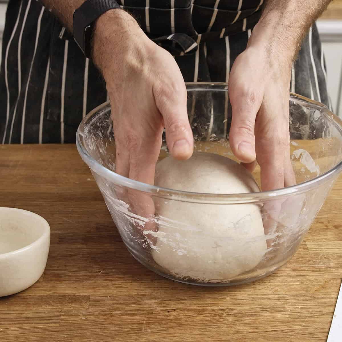 Hands tuck the edges of sourdough under itself inside a clear bowl.