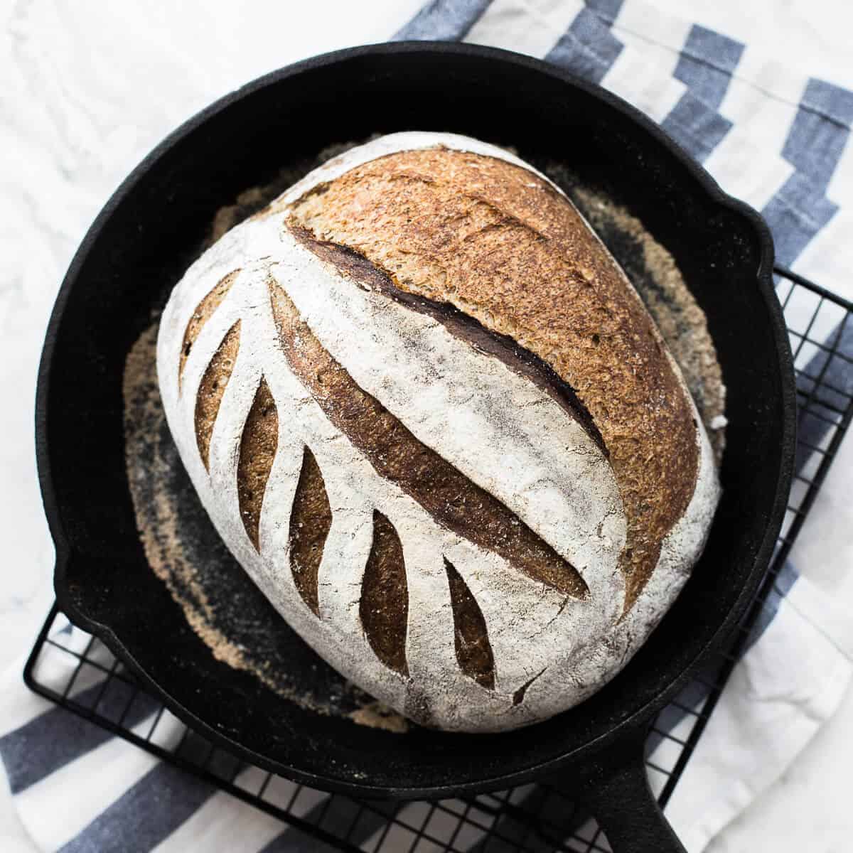 flaxseed meal sourdough in a black skillet on a wire rack and blue and white striped kitchen towel