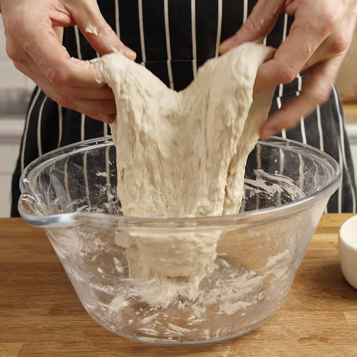 Hands tuck the edges of sourdough under itself inside a clear bowl.