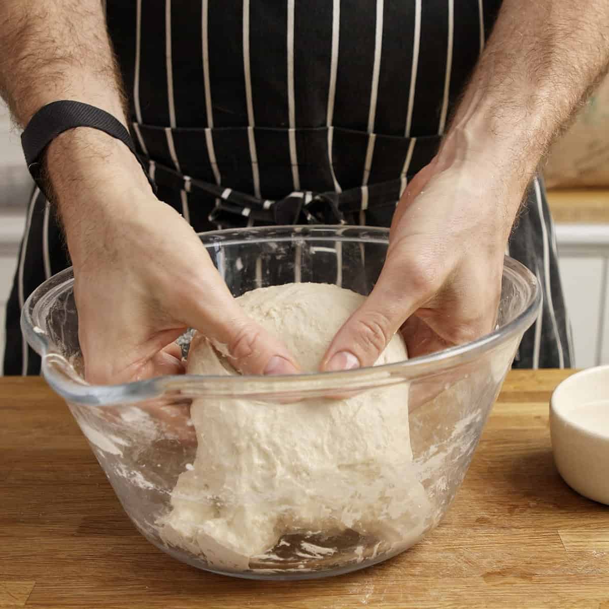Hands tuck the edges of sourdough under itself inside a clear bowl.