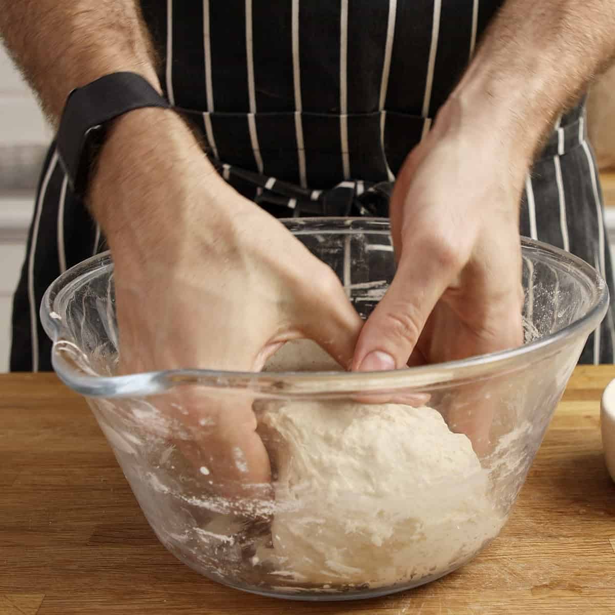 Hands tuck the edges of sourdough under itself inside a clear bowl.