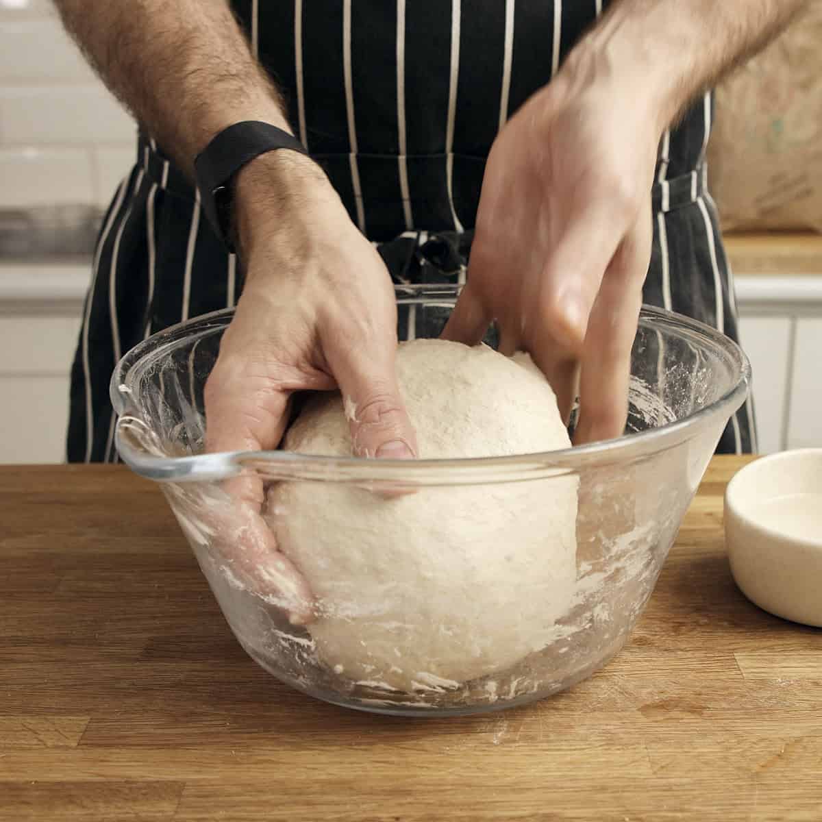 A hand stretches a piece of sourdough upwards inside a clear bowl.