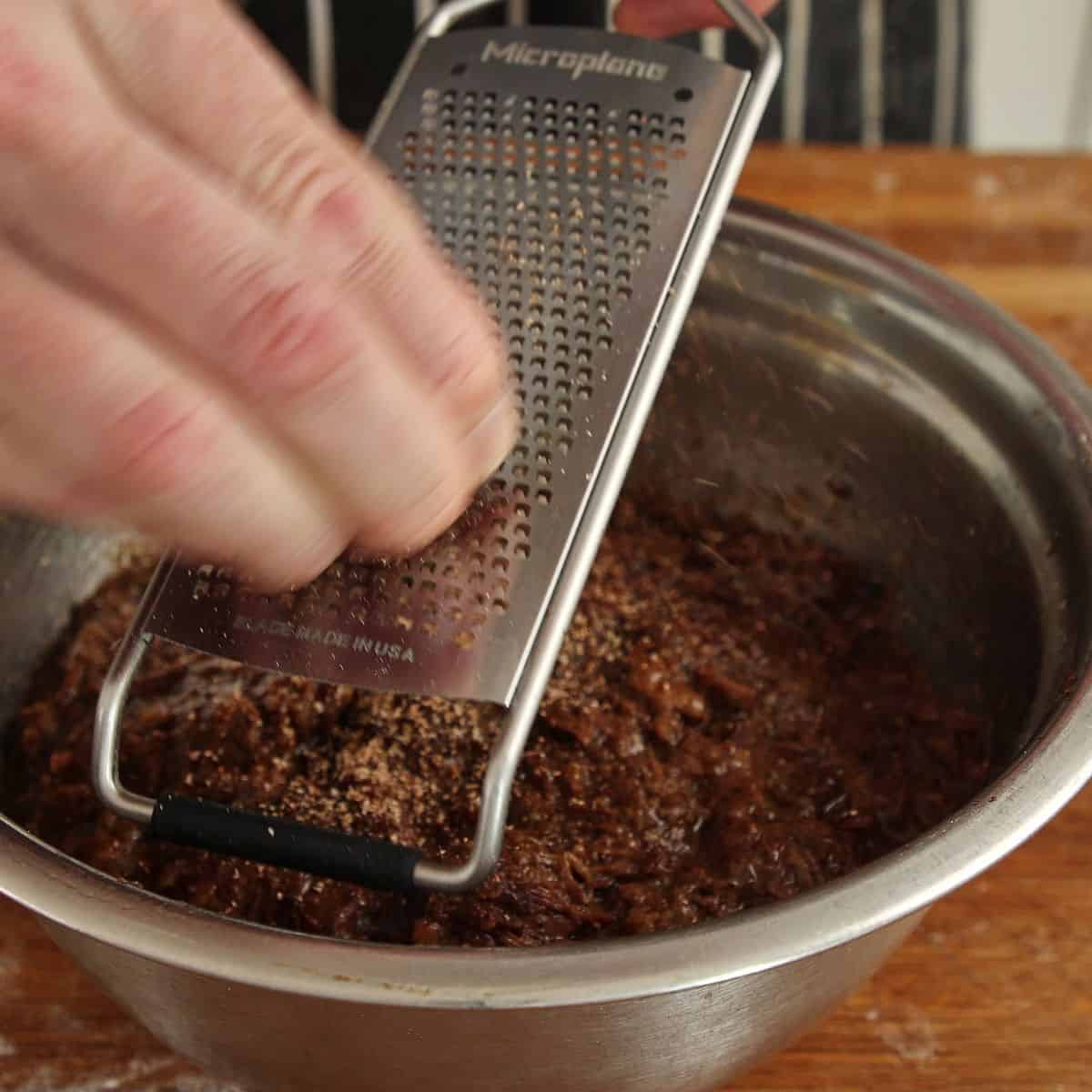 Fresh nutmeg being grated into a metal bowl of dark spiced beef filling using a microplane grater held by hand