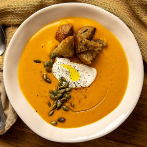 Pumpkin cream soup with crispy croutons, cream and pumpkin seeds in a white bowl.