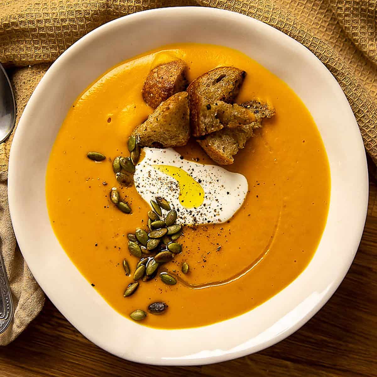 Pumpkin cream soup with crispy croutons, cream and pumpkin seeds in a white bowl.