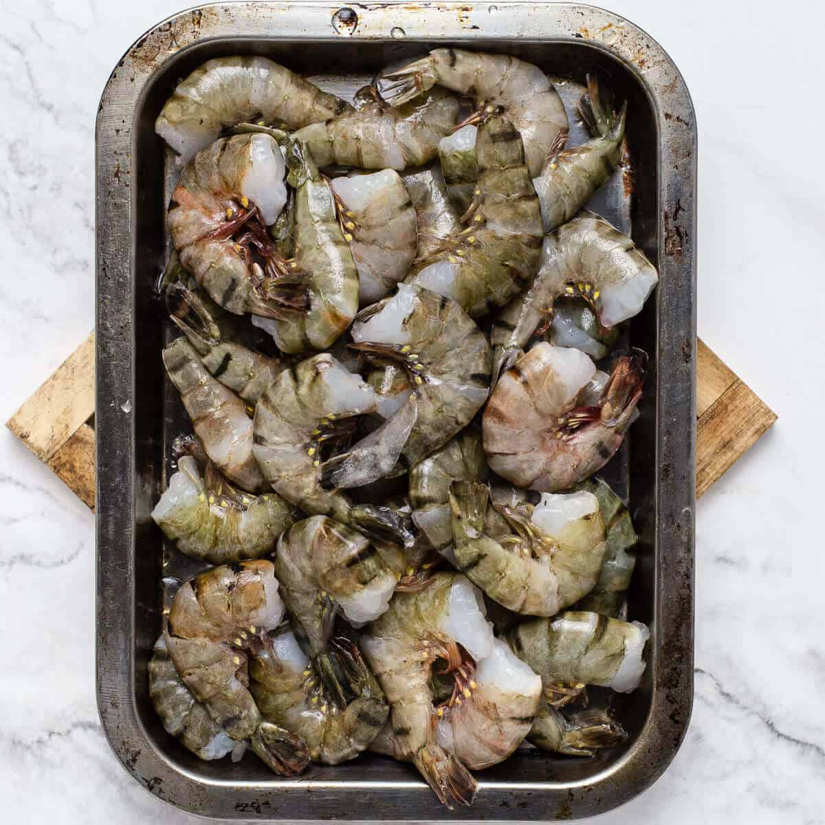 headless raw shrimps on a metal tray on a wooden coaster and marble background