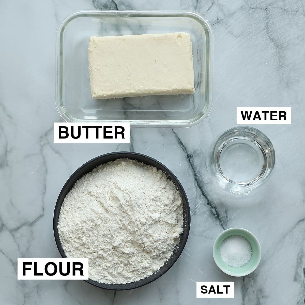 Ingredients for rough puff pastry arranged on white marble — cold cubed butter, flour, salt, ice water in small bowl