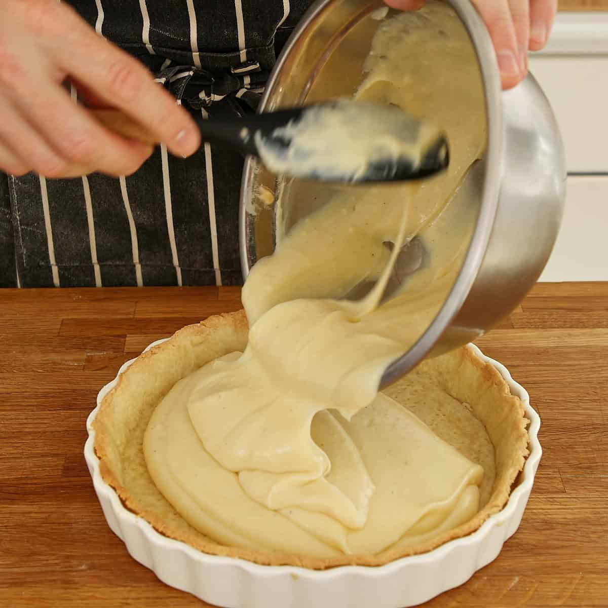 Pastry cream being poured into a baked pie shell in a white ceramic dish.