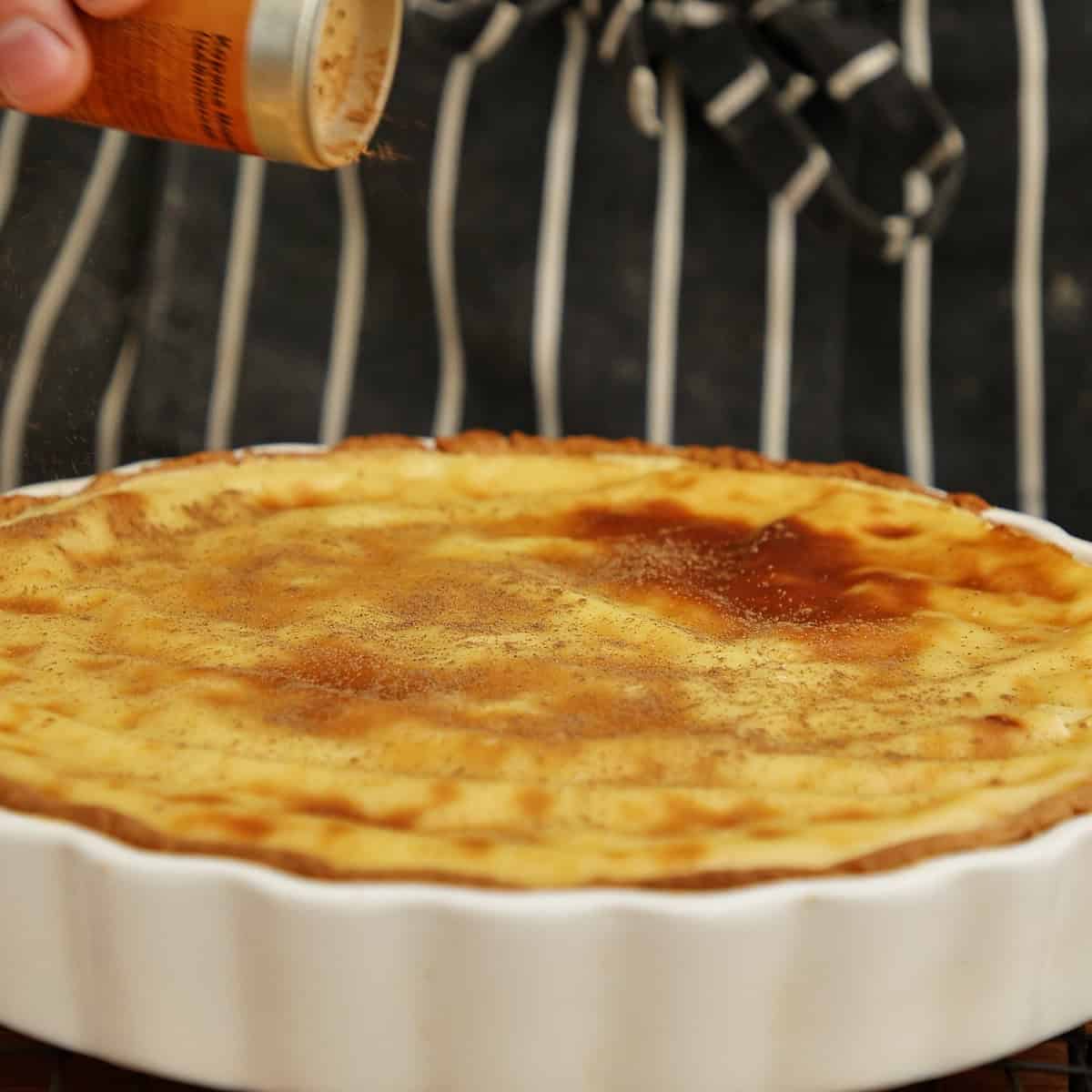 Cinnamon being dusted onto a baked custard tart in a white dish.