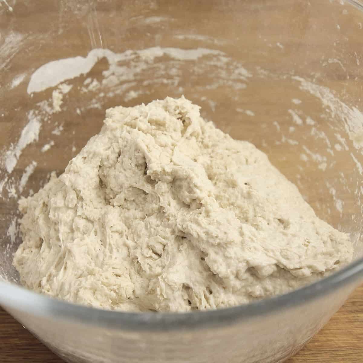 A shaggy sourdough mixture rests in a clear glass bowl.