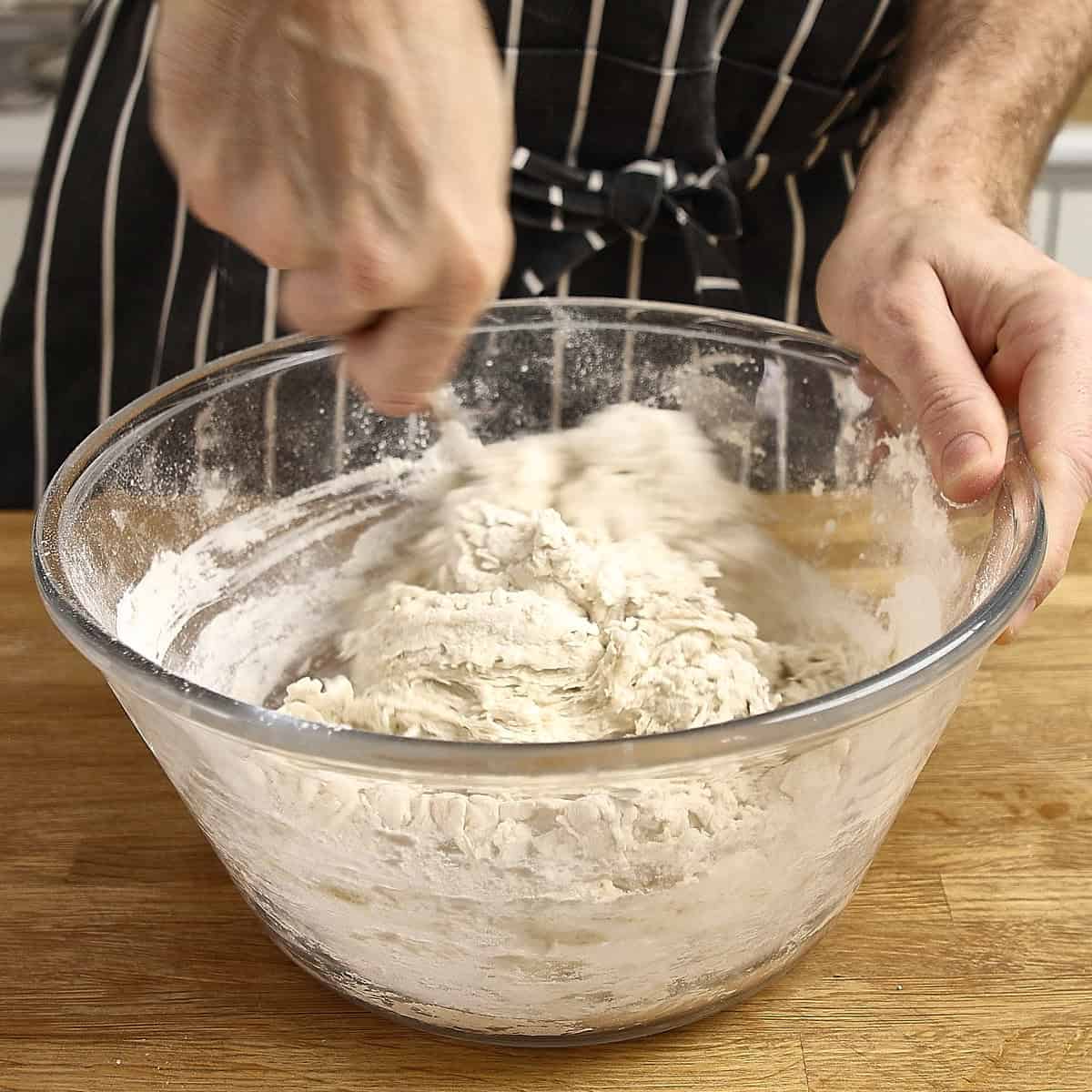 Hands mix a sourdough mixture in a clear glass bowl.