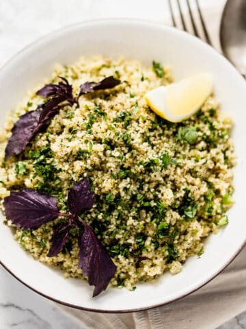 moroccan spiced couscous salad in a white bowl on marble background with purple basil and a wedge of lemon