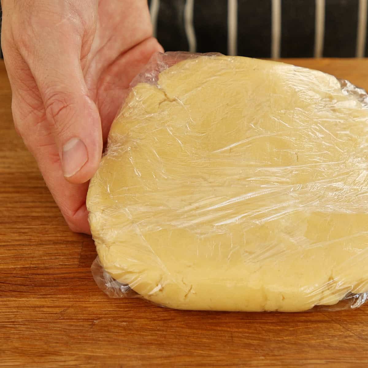 Chilled pastry dough wrapped in plastic film, held by hand on wooden surface.