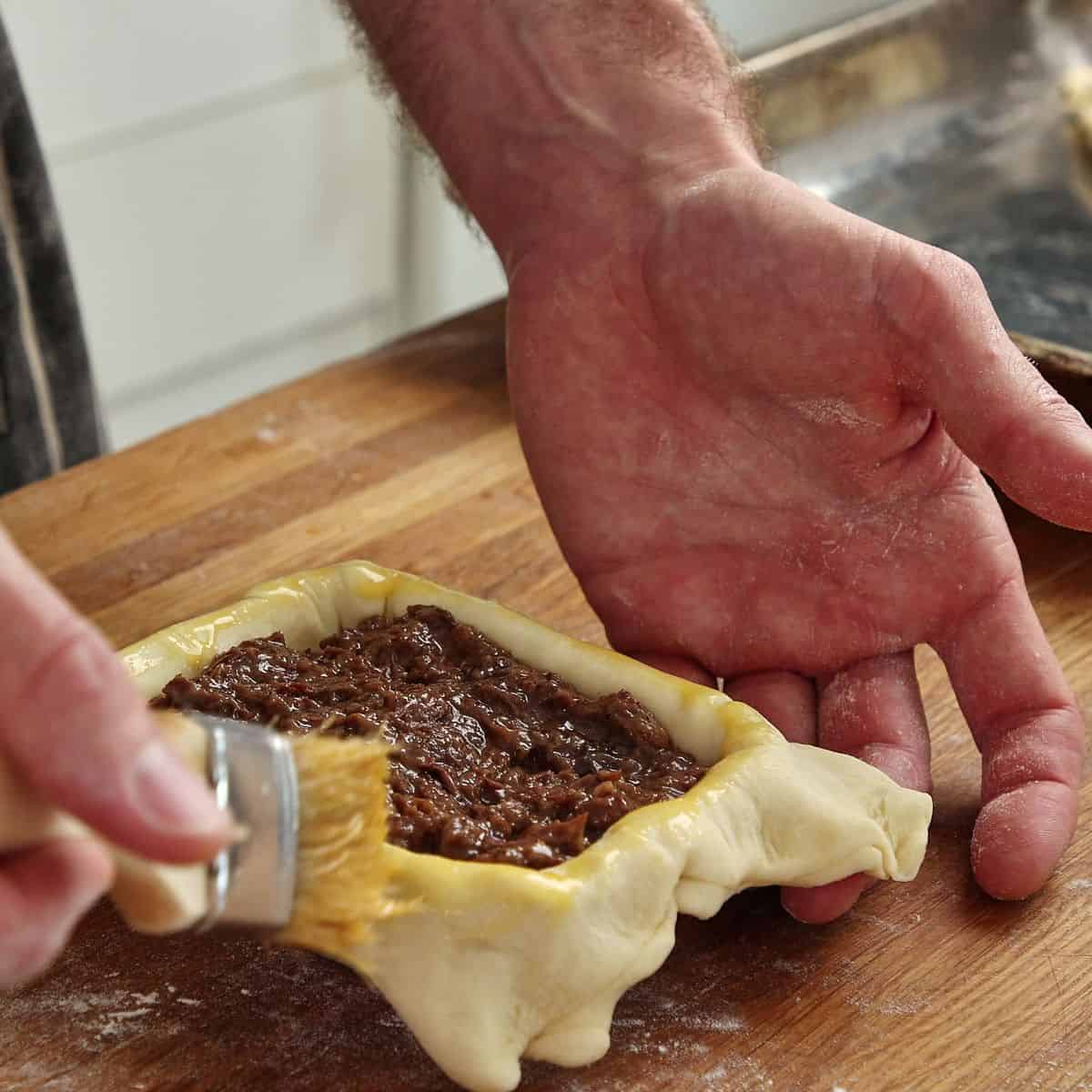 Pastry edges being brushed with egg wash before lid placement, egg wash bowl and brush visible