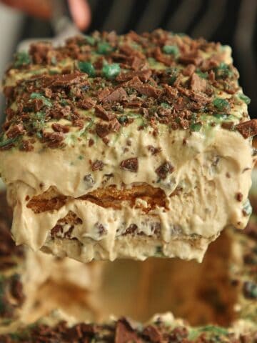 Cross-section of Peppermint Crisp tart on white plate showing distinct layers: caramel mousse, softened coconut Tennis biscuits, mousse, biscuits, and chopped mint chocolate on top, fork cutting through or piece lifted to show interior.