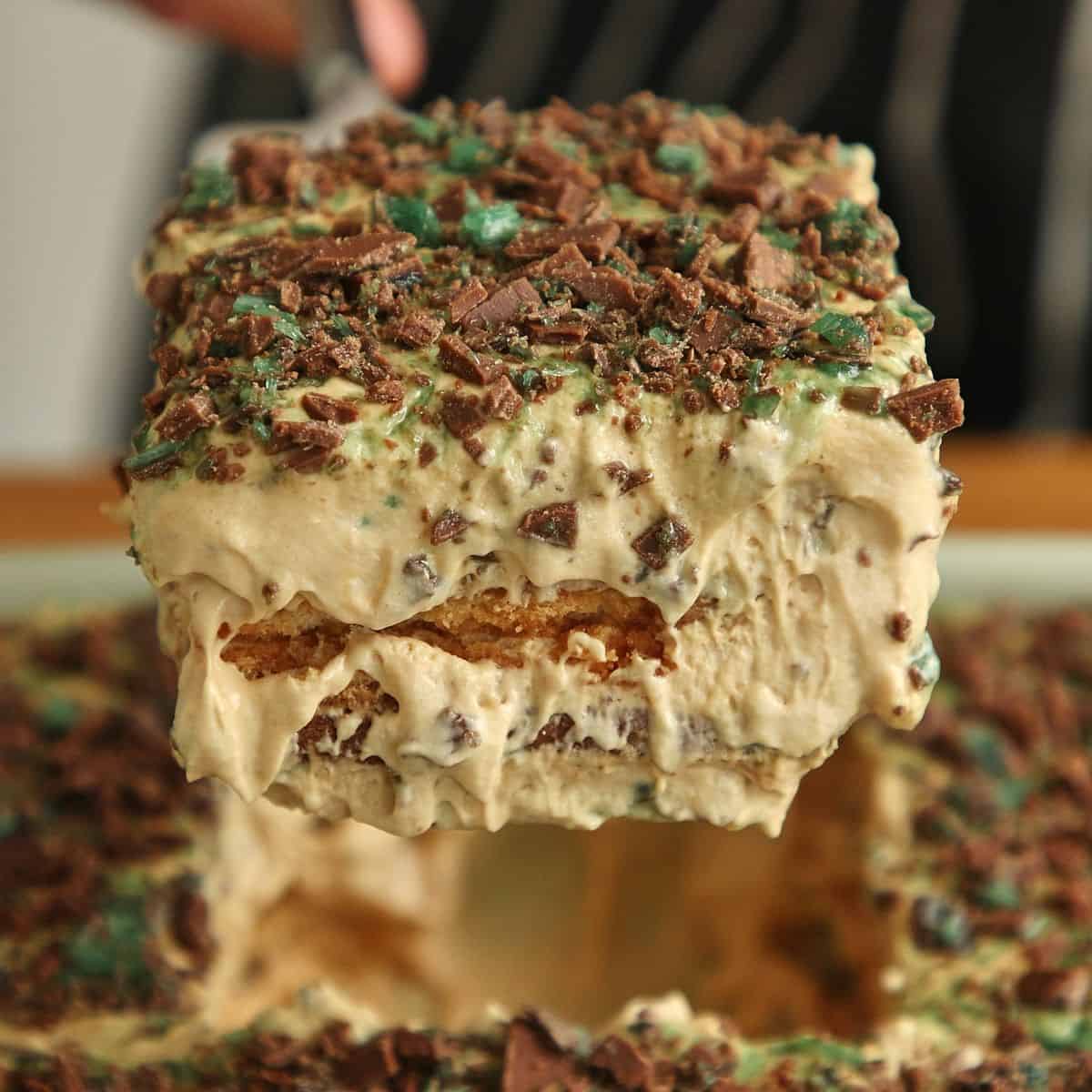 Cross-section of Peppermint Crisp tart on white plate showing distinct layers: caramel mousse, softened coconut Tennis biscuits, mousse, biscuits, and chopped mint chocolate on top, fork cutting through or piece lifted to show interior