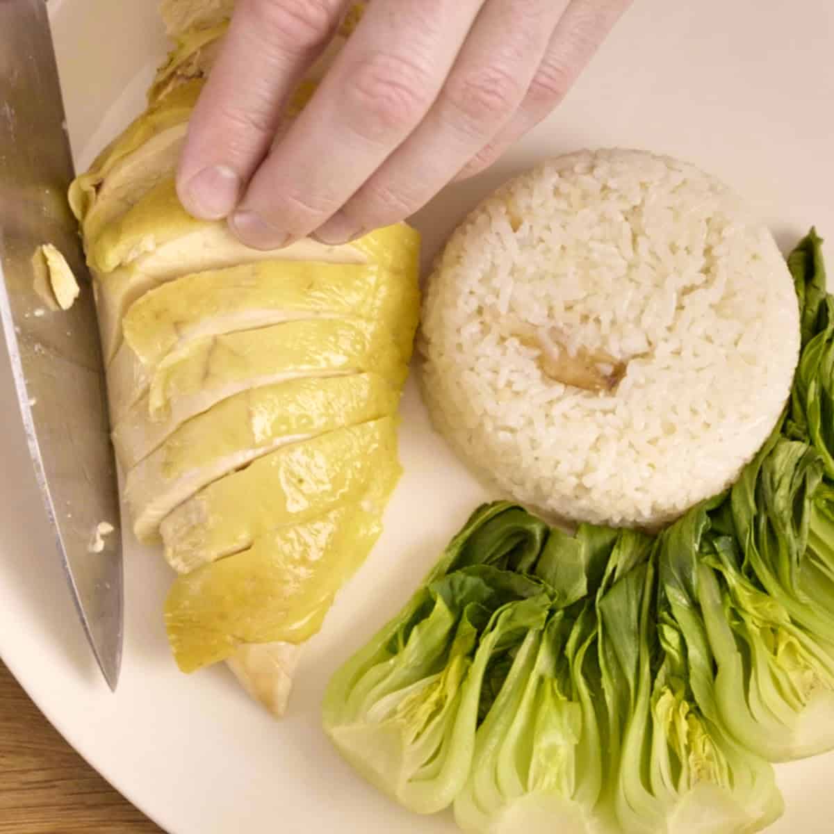 Plating sliced chicken breast onto a white plate with steamed rice and greens.