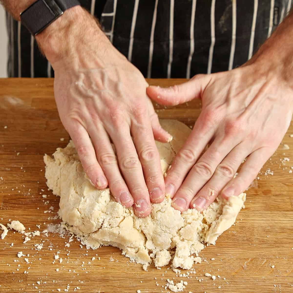Hands pressing crumbly pastry dough together on a wooden board.
