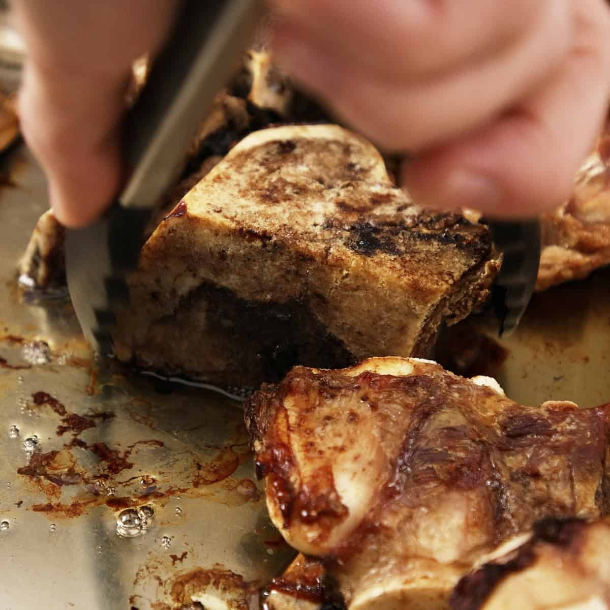 Lifting roasted bones out of roasting tray with tongs.
