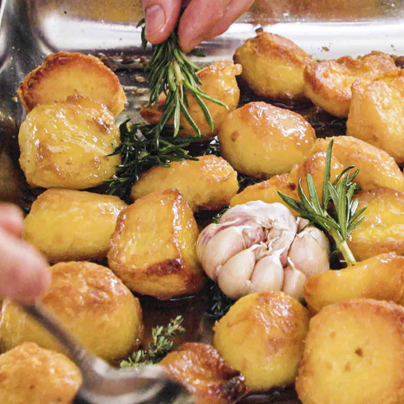 Adding rosemary and garlic to half roasted potatoes on a baking sheet.