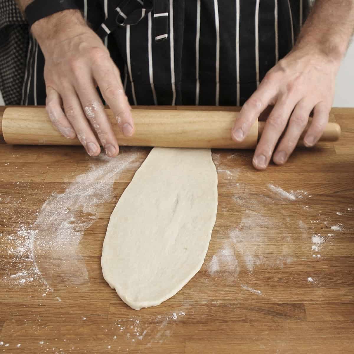 Rolling out naan with rolling pin on floured wooden surface.