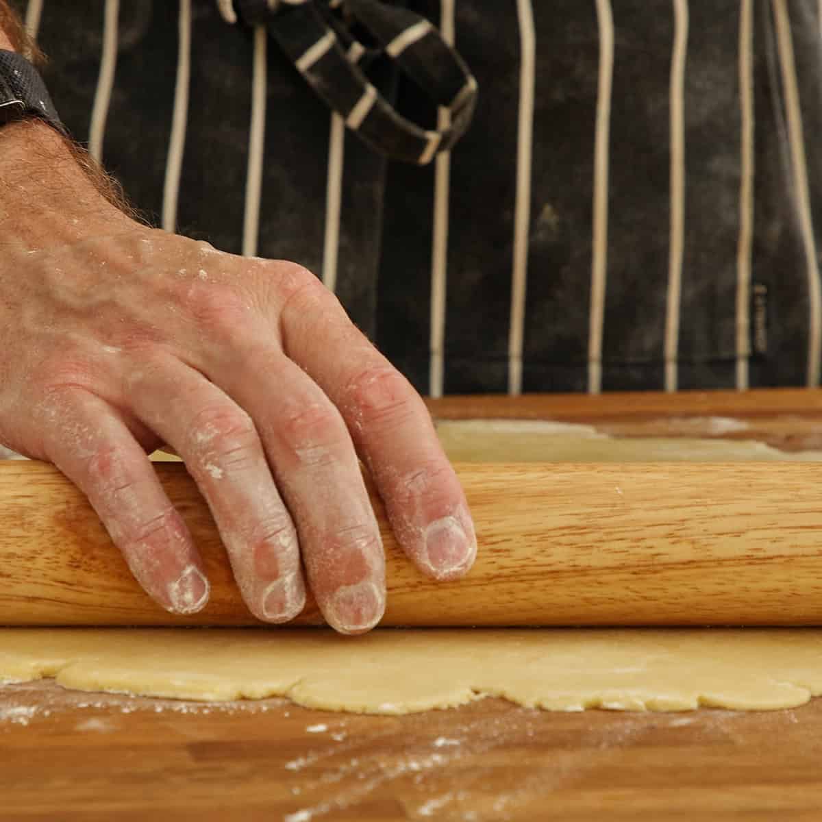 Hand rolling out pale pastry dough with a wooden pin on a floured wooden board.