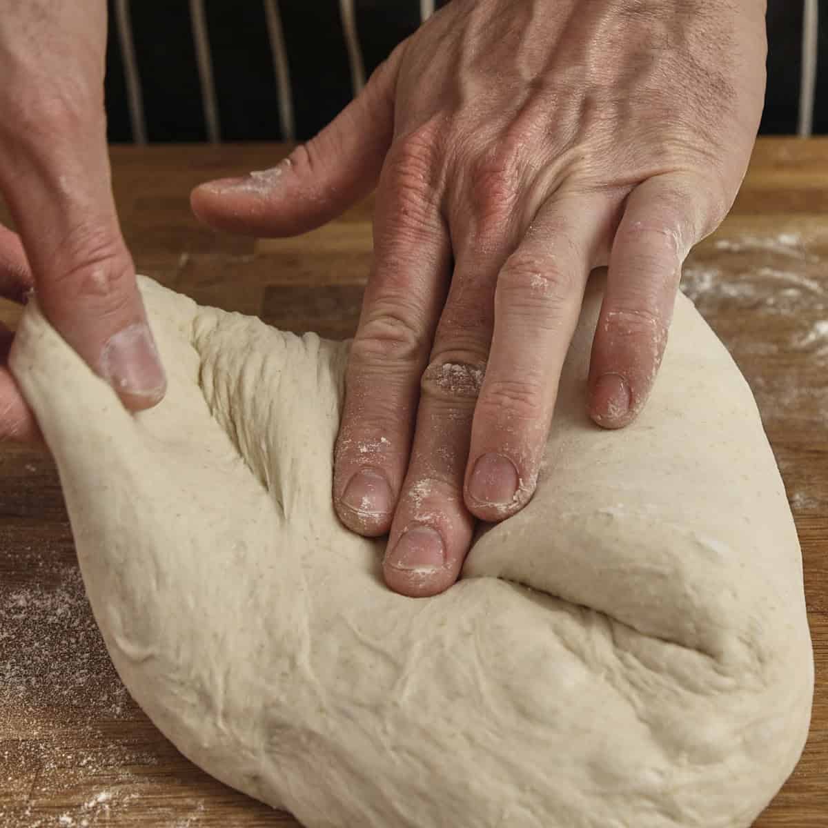 Hands press down on a folded piece of sourdough on a wooden surface.