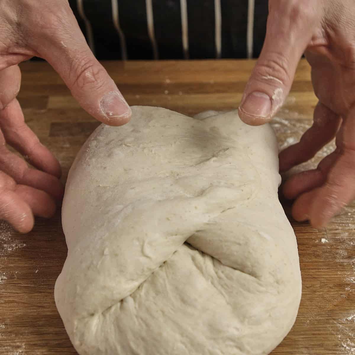 Hands rest next to a folded piece of sourdough on a wooden surface.