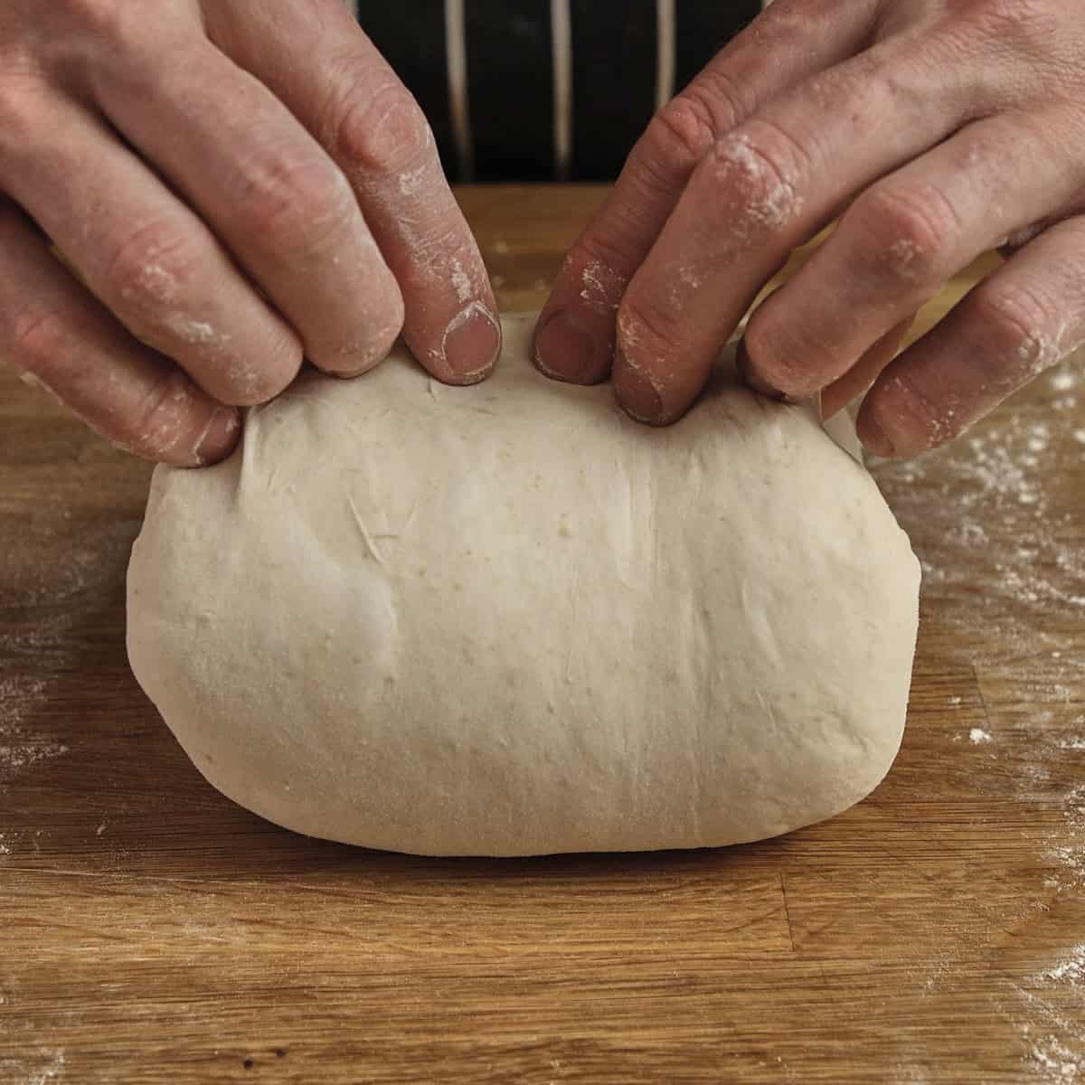 Hands stretch the top edge of a flat piece of sourdough on a wooden surface.