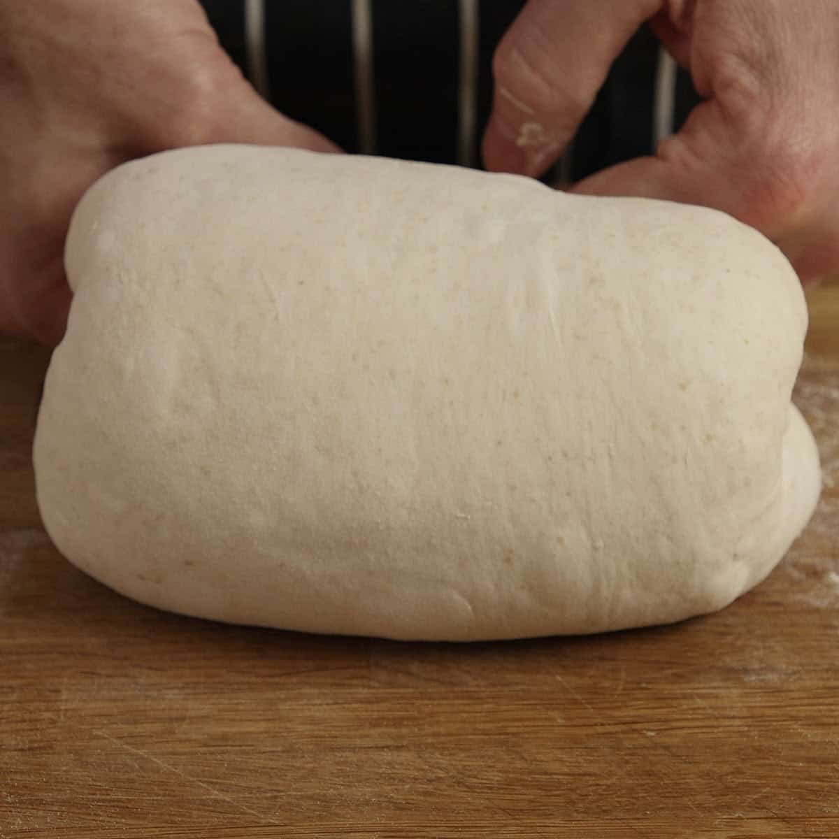Hands shape a smooth loaf of sourdough on a wooden surface.