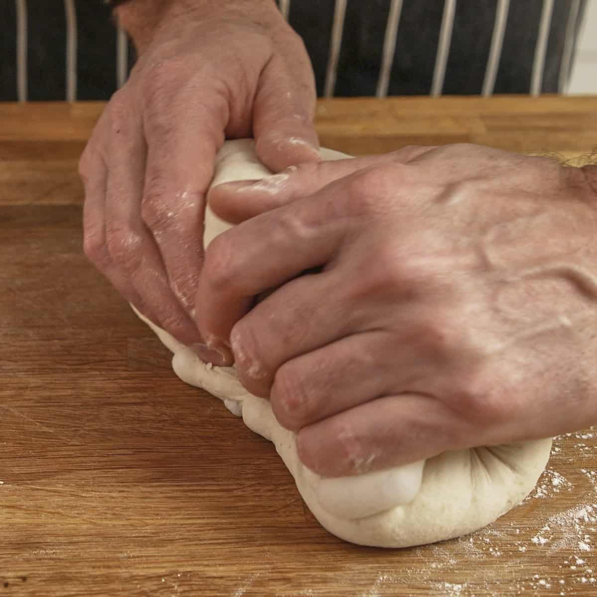 Hands roll a piece of sourdough on a wooden surface.