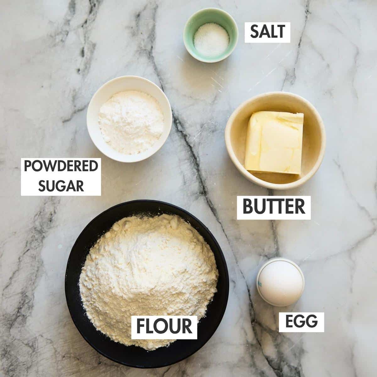 Flour, sugar, salt, butter, and egg arranged in separate bowls on marble.
